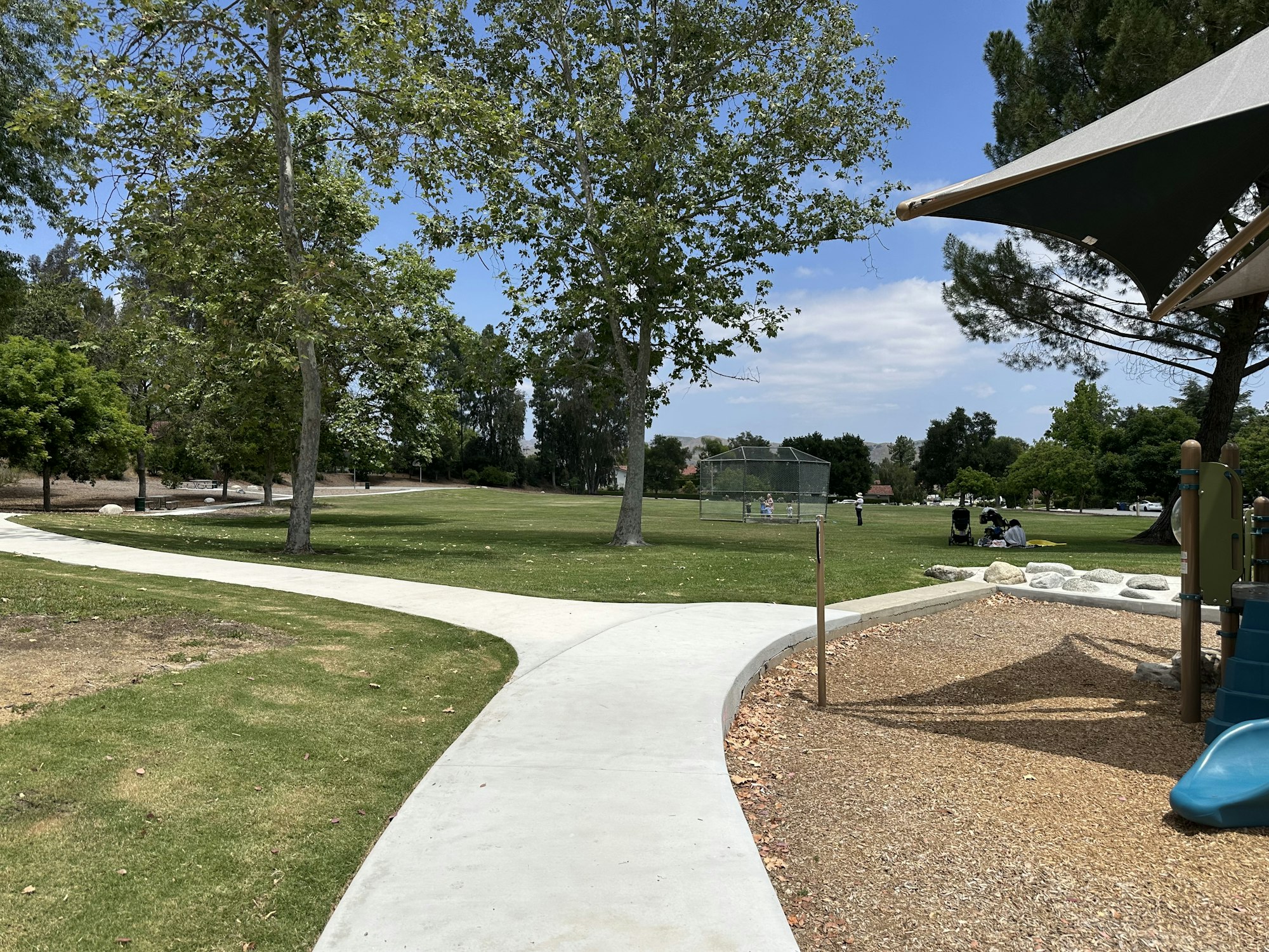 A sunny park scene with walking paths, trees, a playground, and a baseball area where people are enjoying outdoor activities.