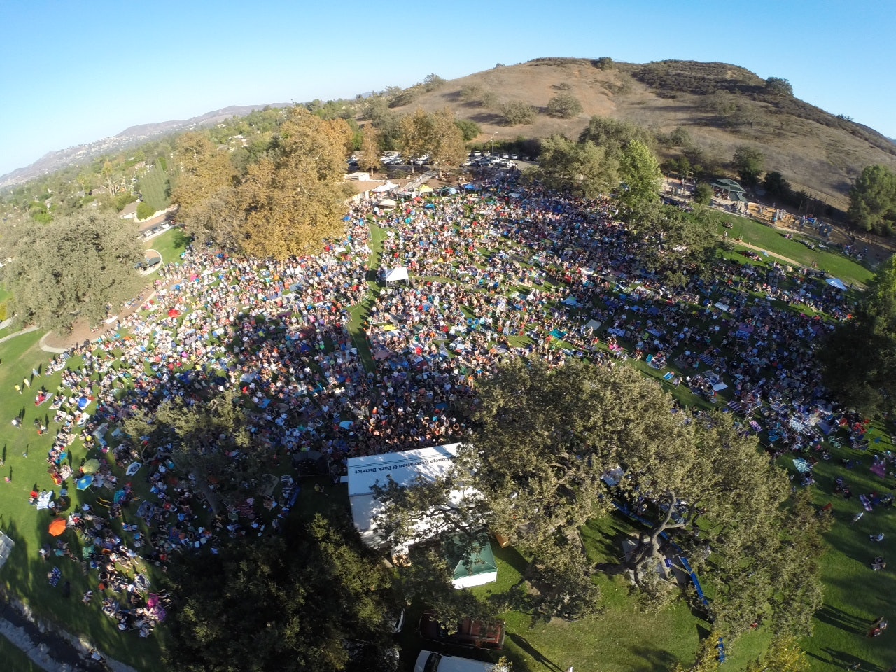 Aerial view of a large crowd gathered in a park for an outdoor event, surrounded by trees and hills.