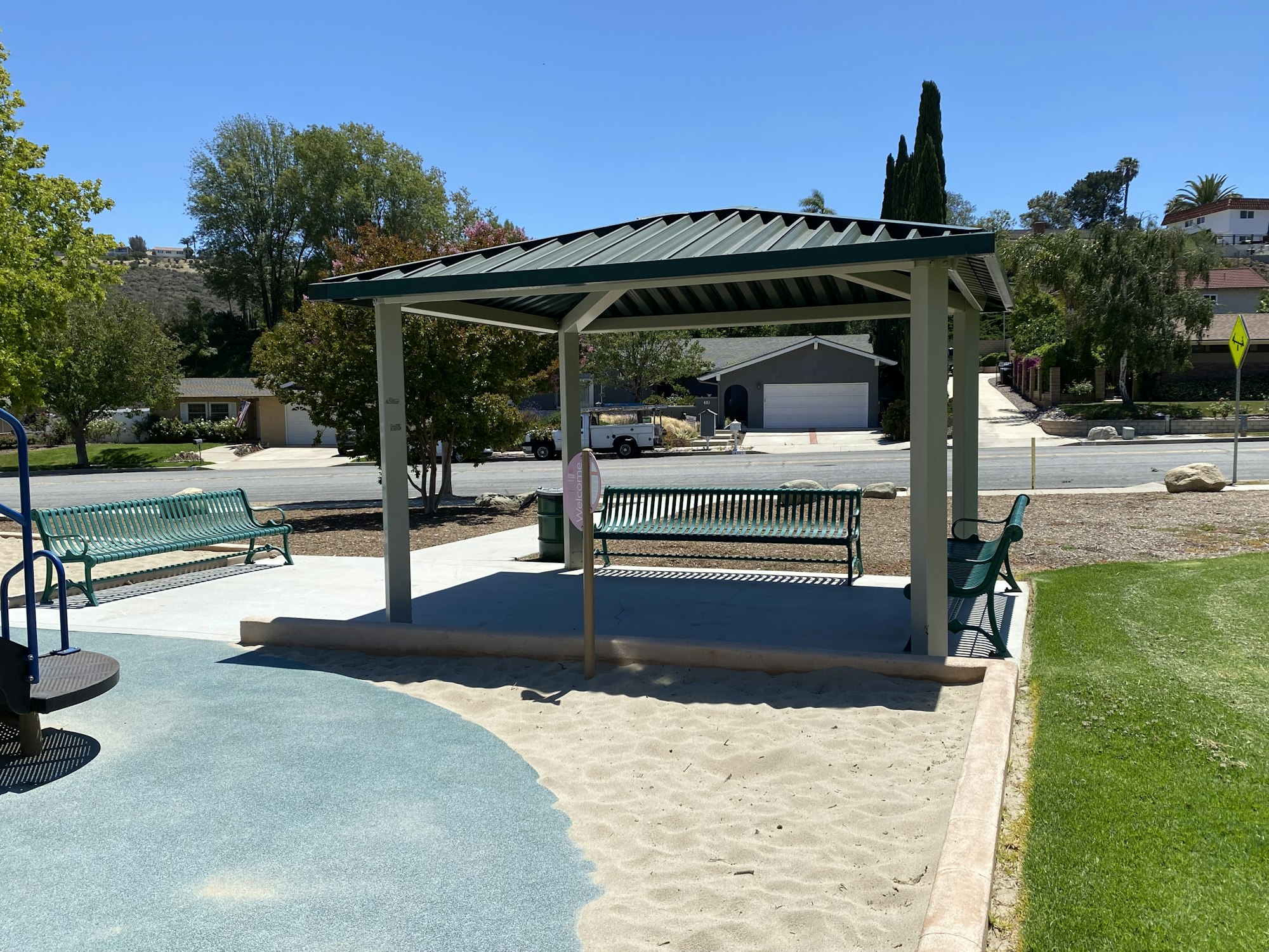 A playground with a canopy, sand area, benches, and a green grass lawn, surrounded by trees and houses in the background.
