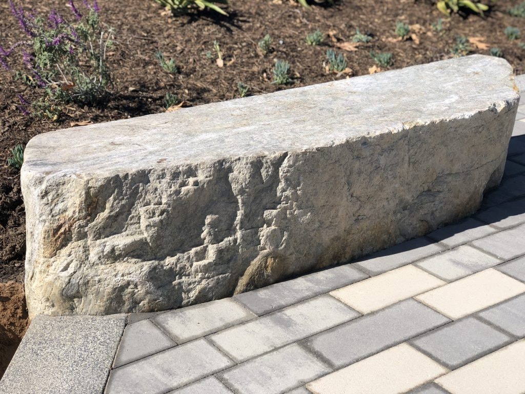 A stone bench on a tiled path with soil and plants in the background.