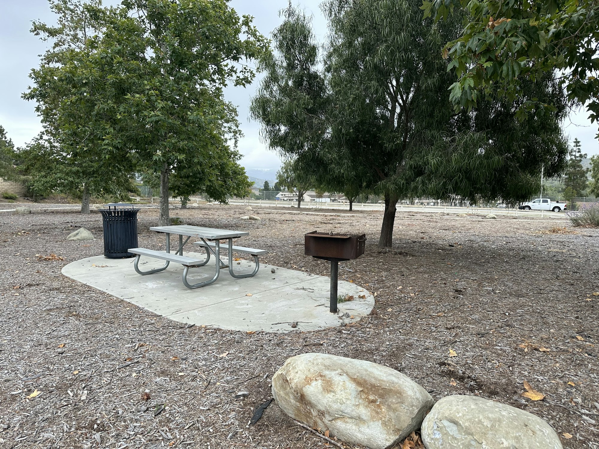 A picnic area with a metal table, grill, trash can, and trees in a gravel park setting under a cloudy sky.