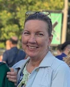A person smiling outdoors with people and greenery in the background.