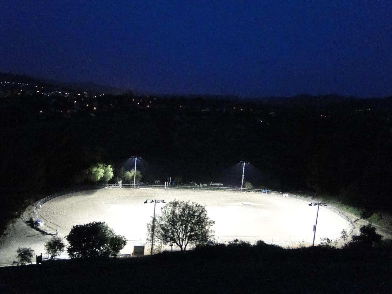 A well-lit empty outdoor arena surrounded by trees at night, with city lights in the background.