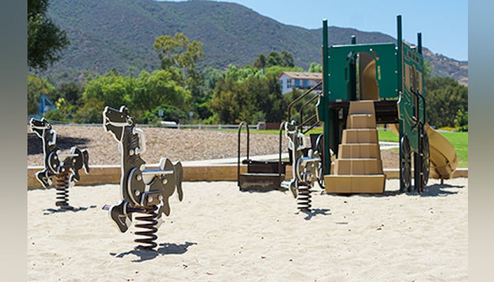 A playground with spring riders and a jungle gym with slides and stairs, set on a sandy area with hills in the background.