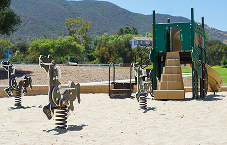 A playground with spring riders and a jungle gym with slides and stairs, set on a sandy area with hills in the background.
