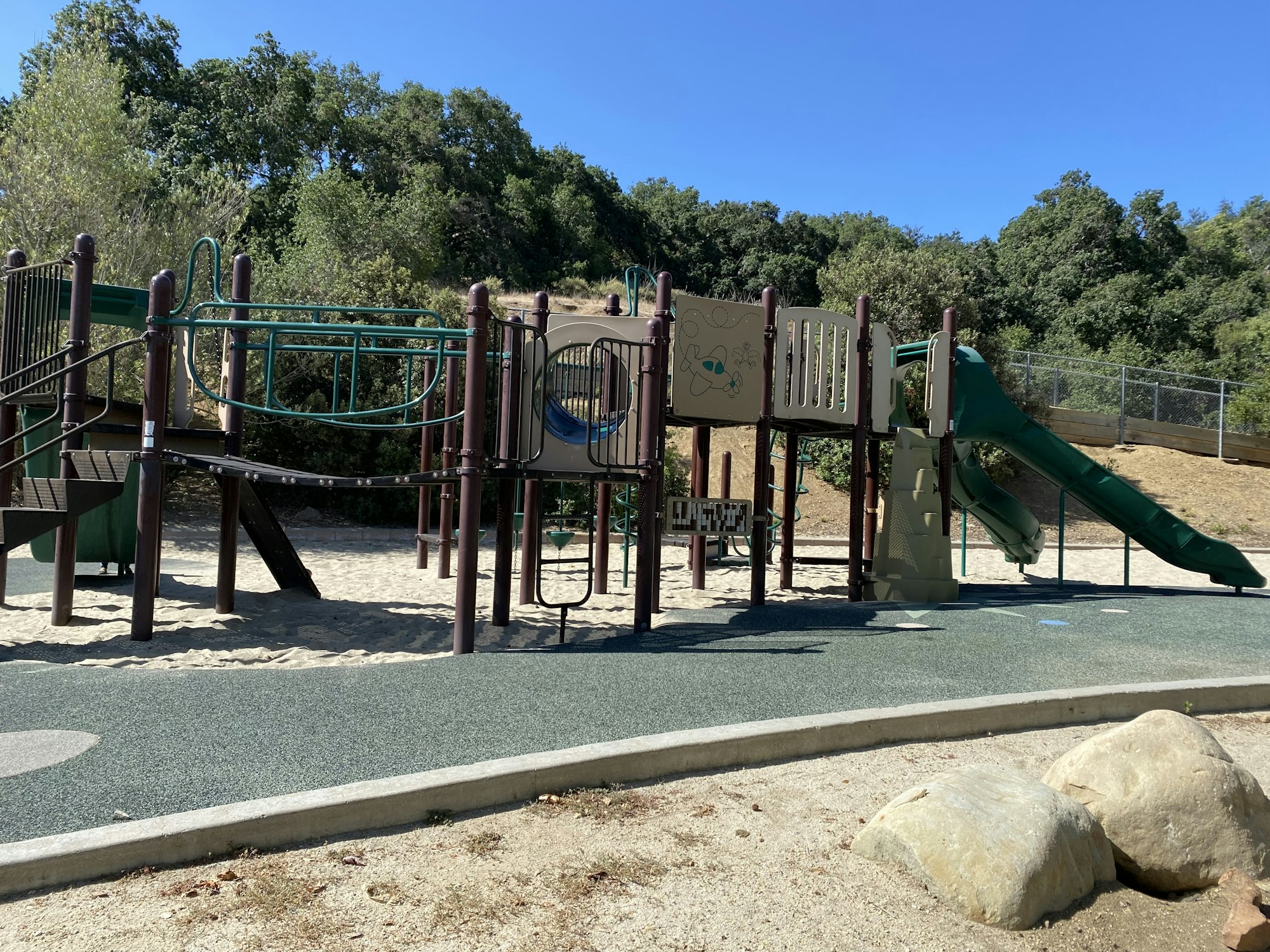 A playground with climbing structures, slides, and a sandy ground surrounded by trees and rocks.