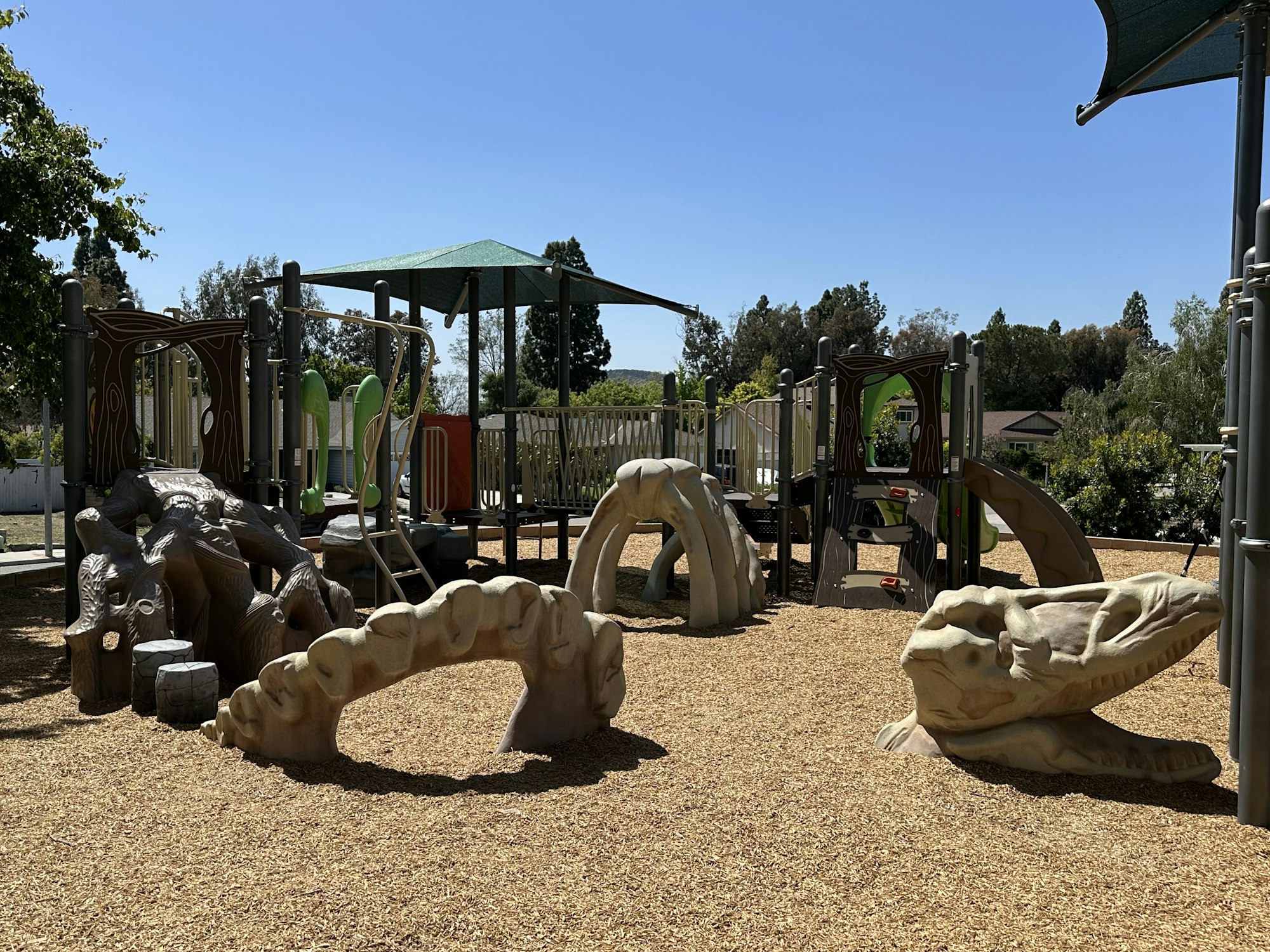 Children's playground with dinosaur-themed structures and slides, surrounded by wood chips under a clear sky.