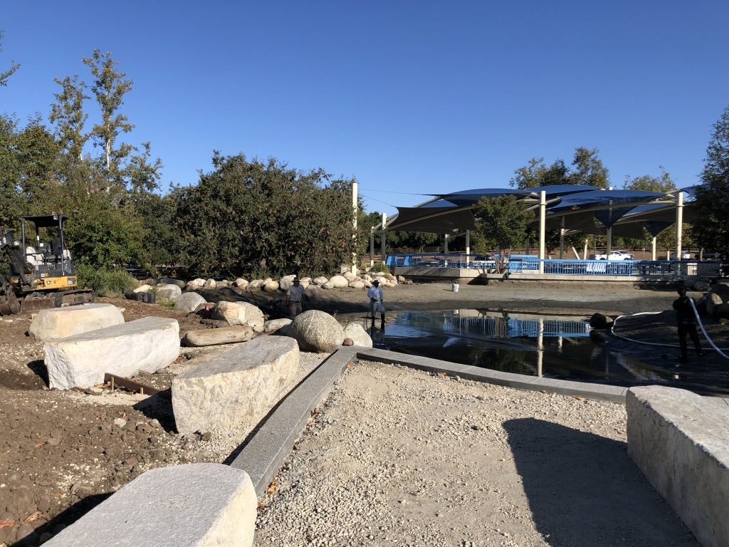Outdoor area under construction with large rocks, a small pond, workers, and a structure with blue shade covers in the background.