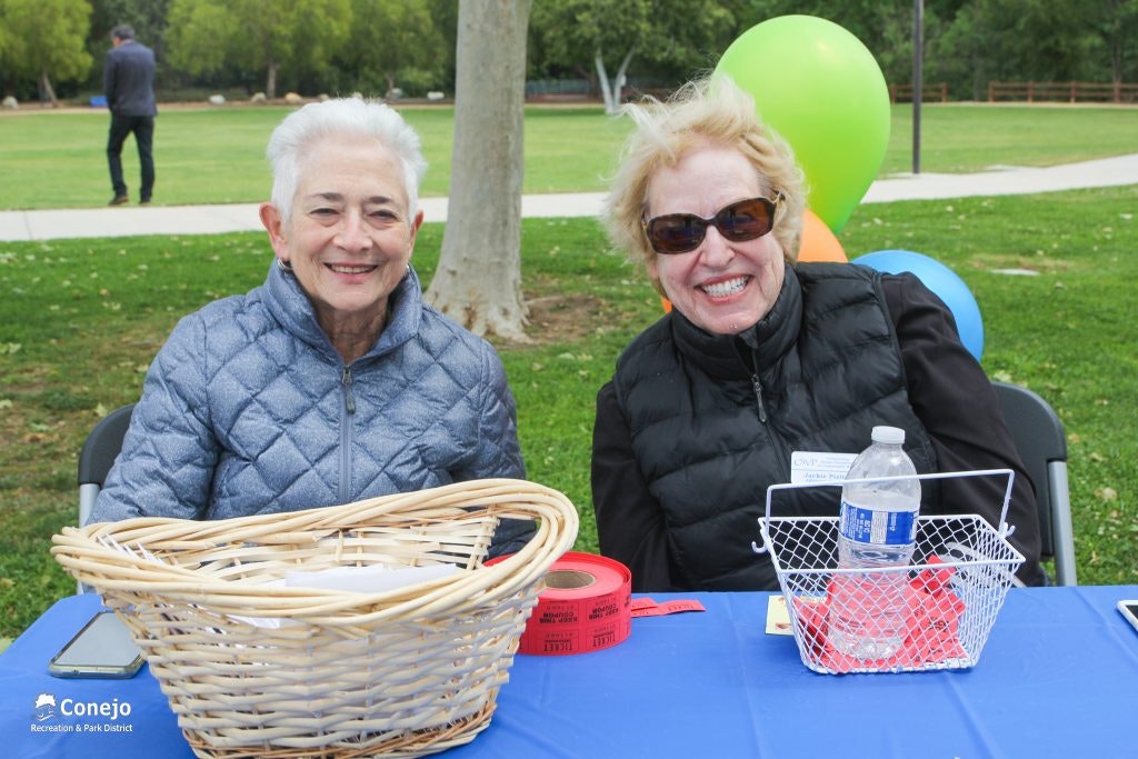 Two people sitting at a table outdoors with baskets, a water bottle, and colorful balloons in the background.