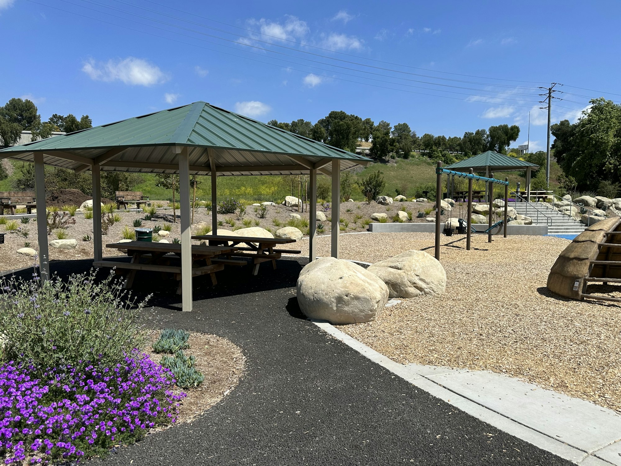Outdoor park with picnic shelters, playground equipment, large rocks, and vibrant flowers under a clear blue sky.