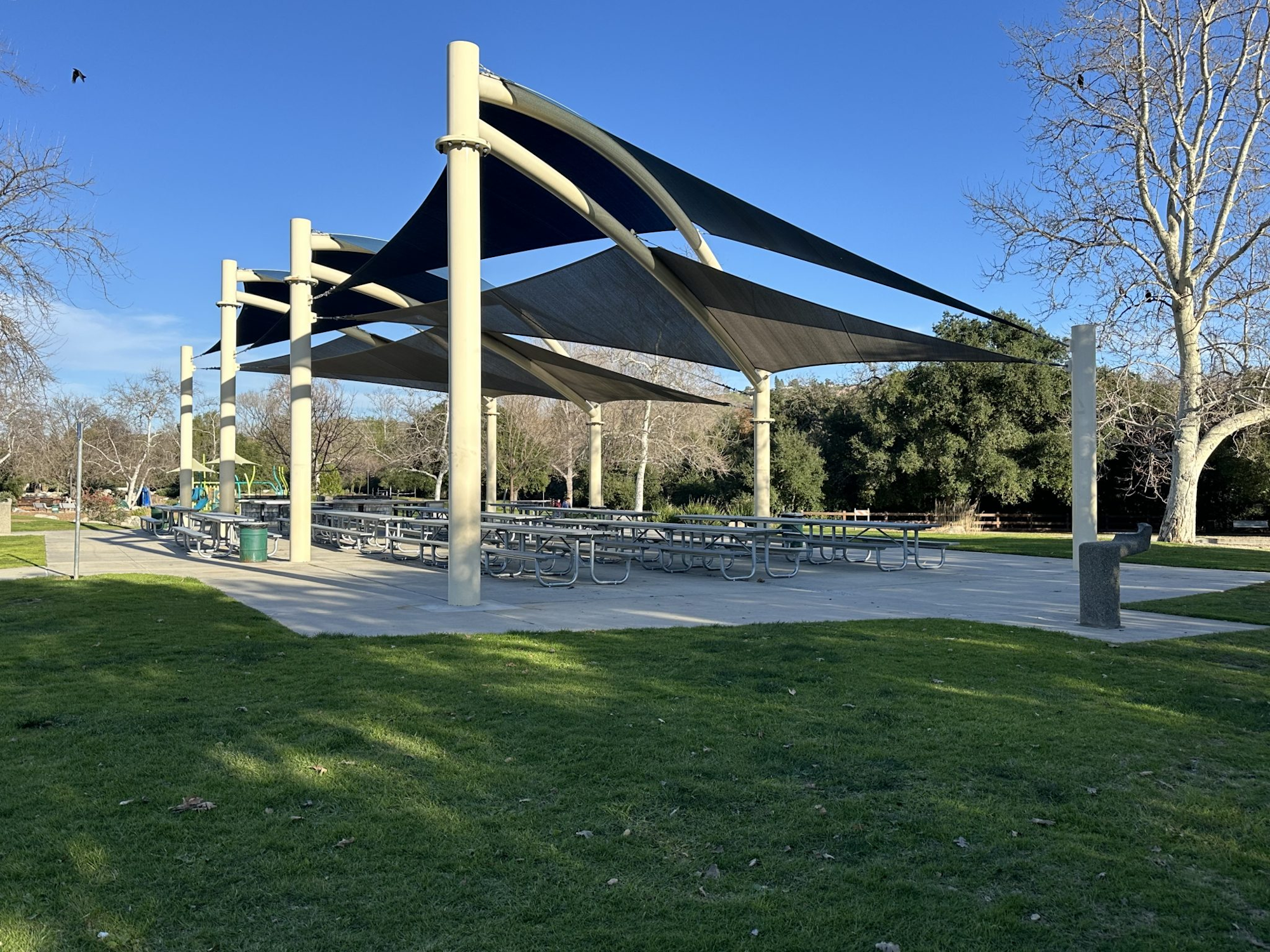 A park pavilion with picnic tables and shade structures, surrounded by grass and trees.