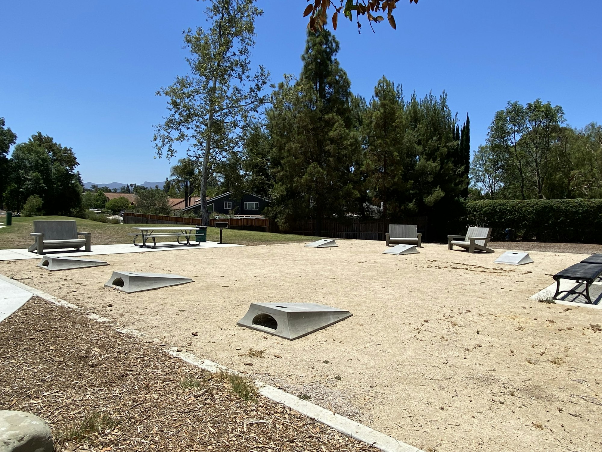 A sandy park area with benches, trees, and cornhole boards under a clear blue sky.