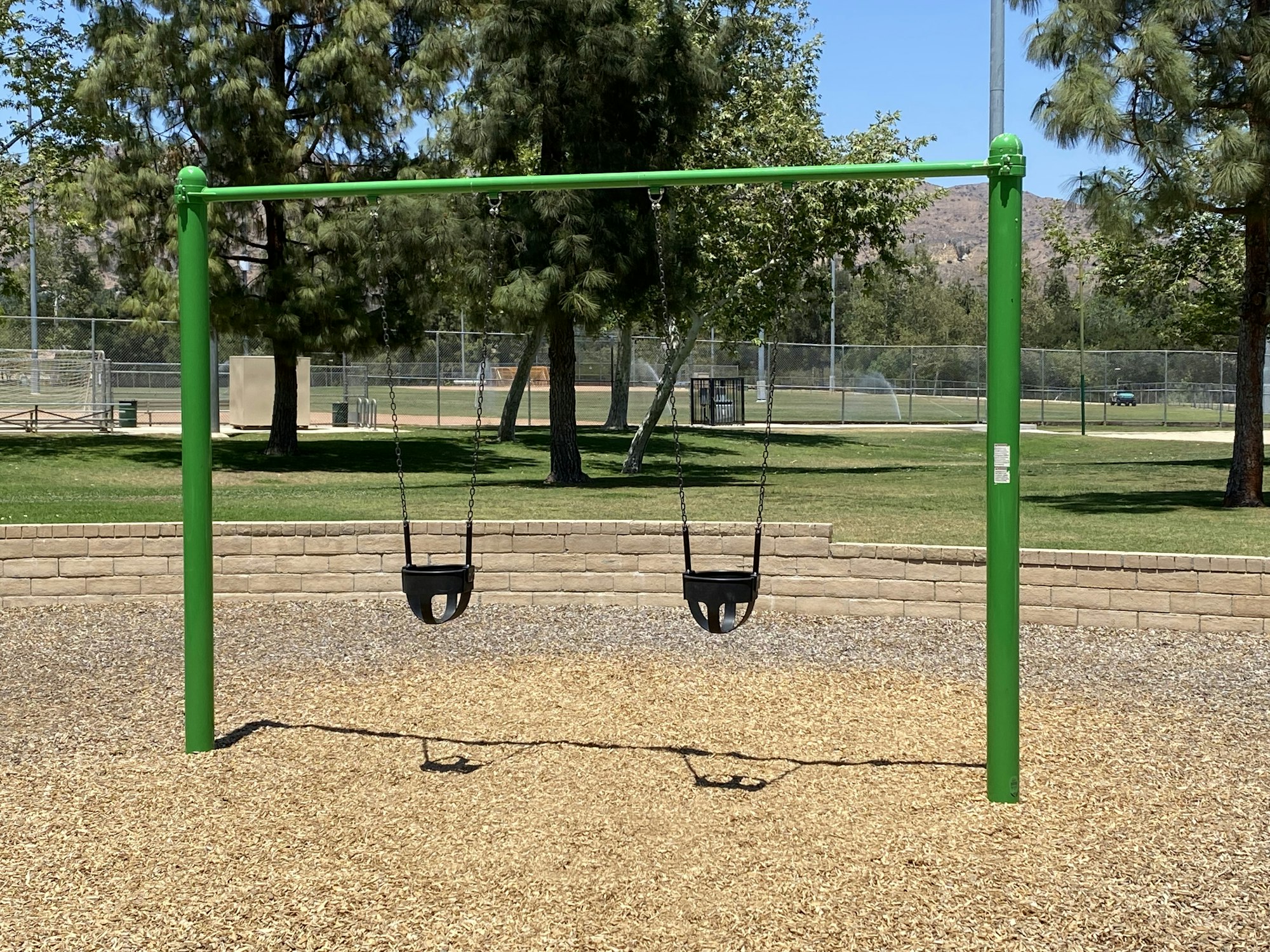 Two empty children's swings on a green frame, set in a playground with trees and a clear sky.