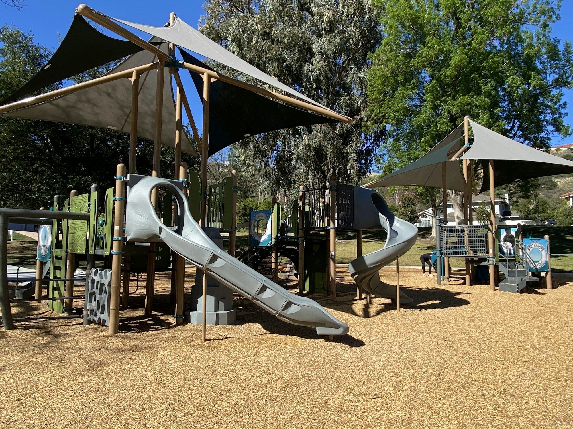 Outdoor playground with slides, climbing structures, and shade canopies surrounded by wood chips.