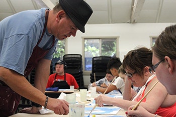 People in a room painting at a table with one person standing and assisting others.