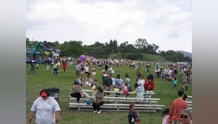 A crowded outdoor event with people on a grassy field, inflatables in the background, and benches filled with guests.