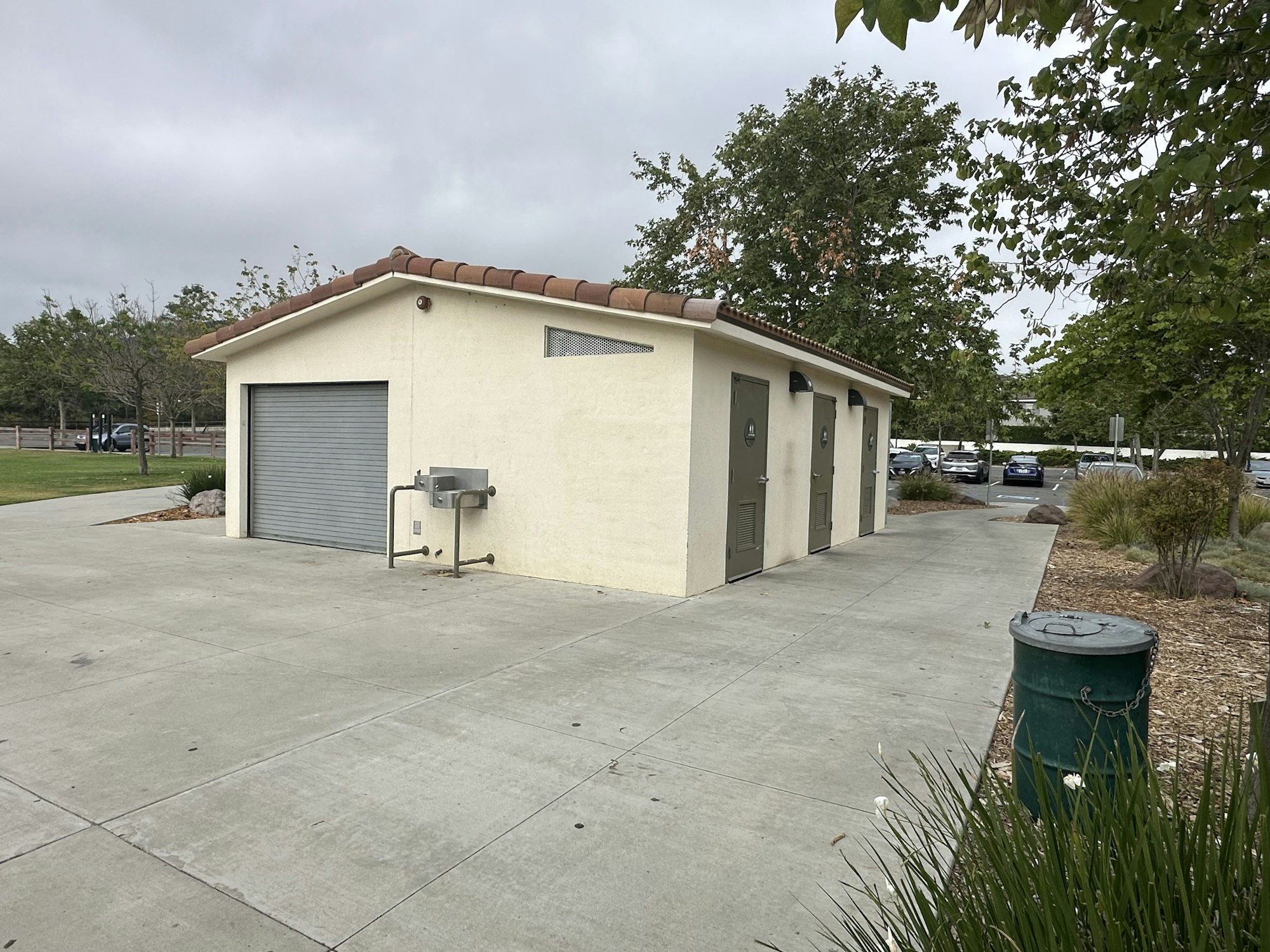 The image shows a small public restroom building with a sloped roof, located in a grassy area with trees and parking nearby.