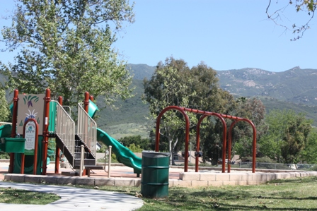 A playground with slides and swings, surrounded by trees and mountains in the background.