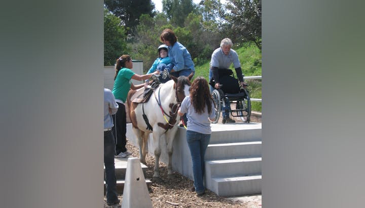 A person on a horse being assisted by multiple individuals. Another person in a wheelchair nearby.