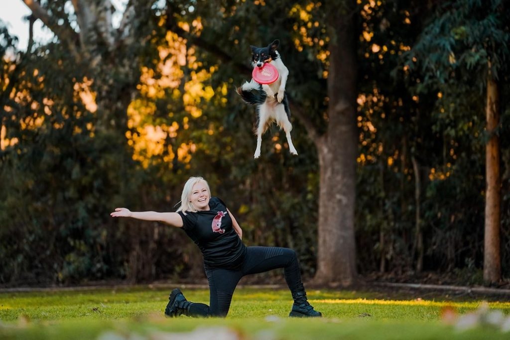 A person kneeling on grass, arms outstretched, as a dog leaps to catch a red frisbee mid-air in a wooded area.