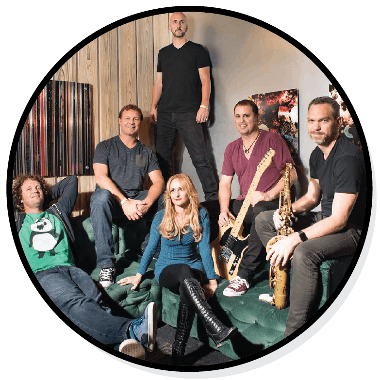 A group of six people posing together in a room, two holding musical instruments.