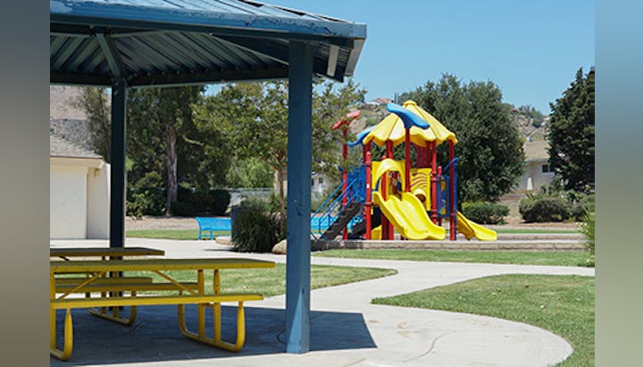 A playground with slides and climbing structures, a covered picnic area, trees, and a clear blue sky.