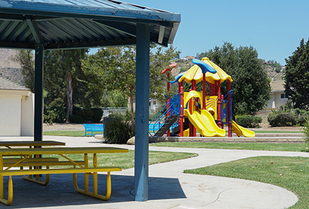 A playground with slides and climbing structures, a covered picnic area, trees, and a clear blue sky.