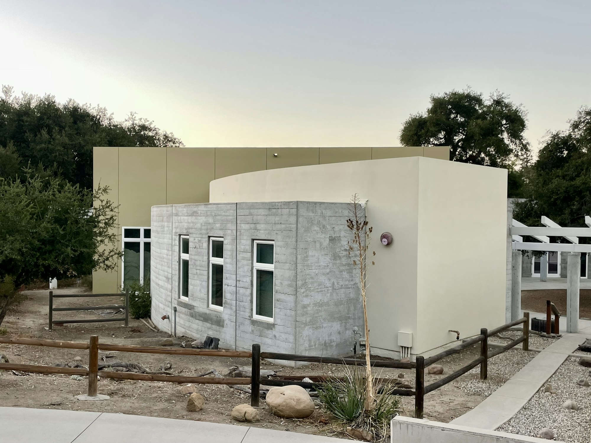 A modern building with a curved concrete wall, large windows, surrounded by sparse vegetation and wooden fencing.