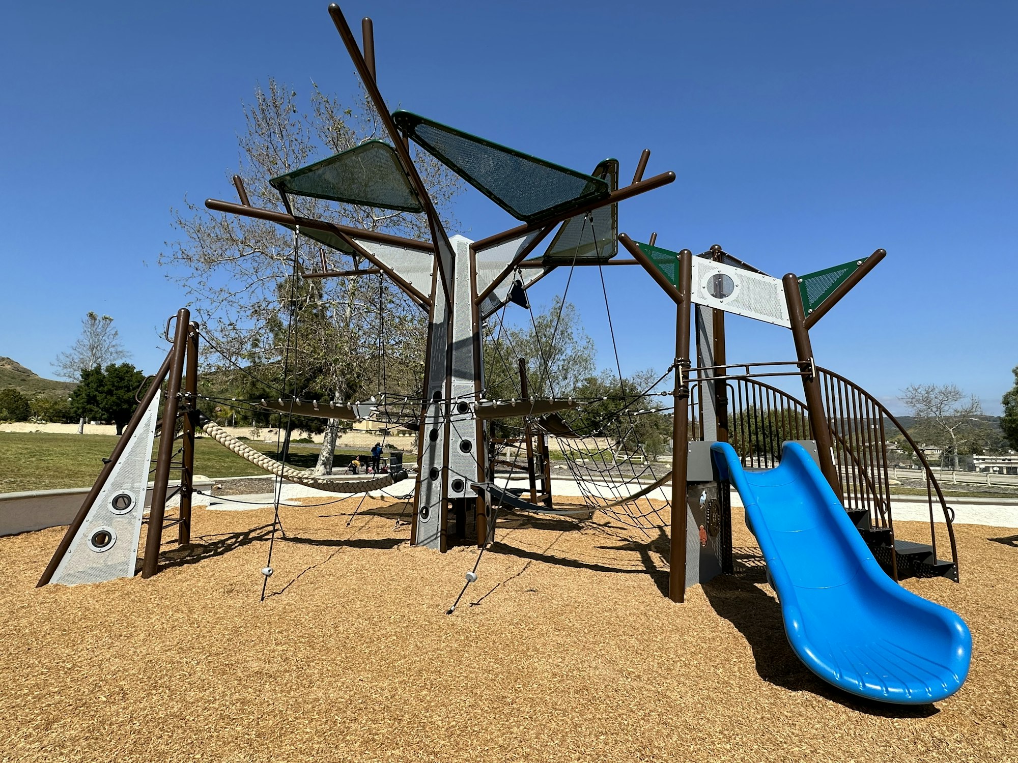 Playground structure with ropes, climbing elements, and a blue slide on wood chips, set in a park area.