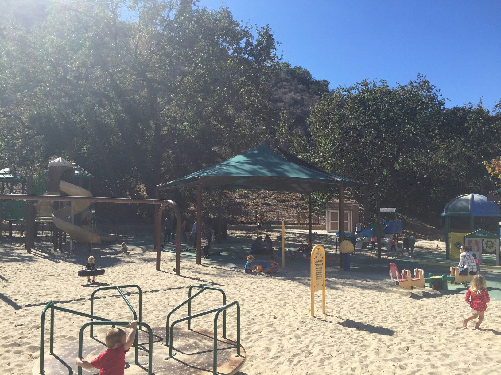 A sunny playground with kids playing on equipment, a slide, and swings. Trees surround the area.