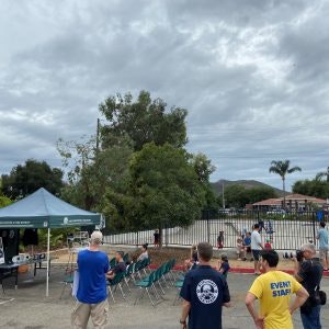 Outdoor gathering with people standing and seated, some wearing "Event Staff" shirts, near a canopy and equipment. Cloudy sky above.