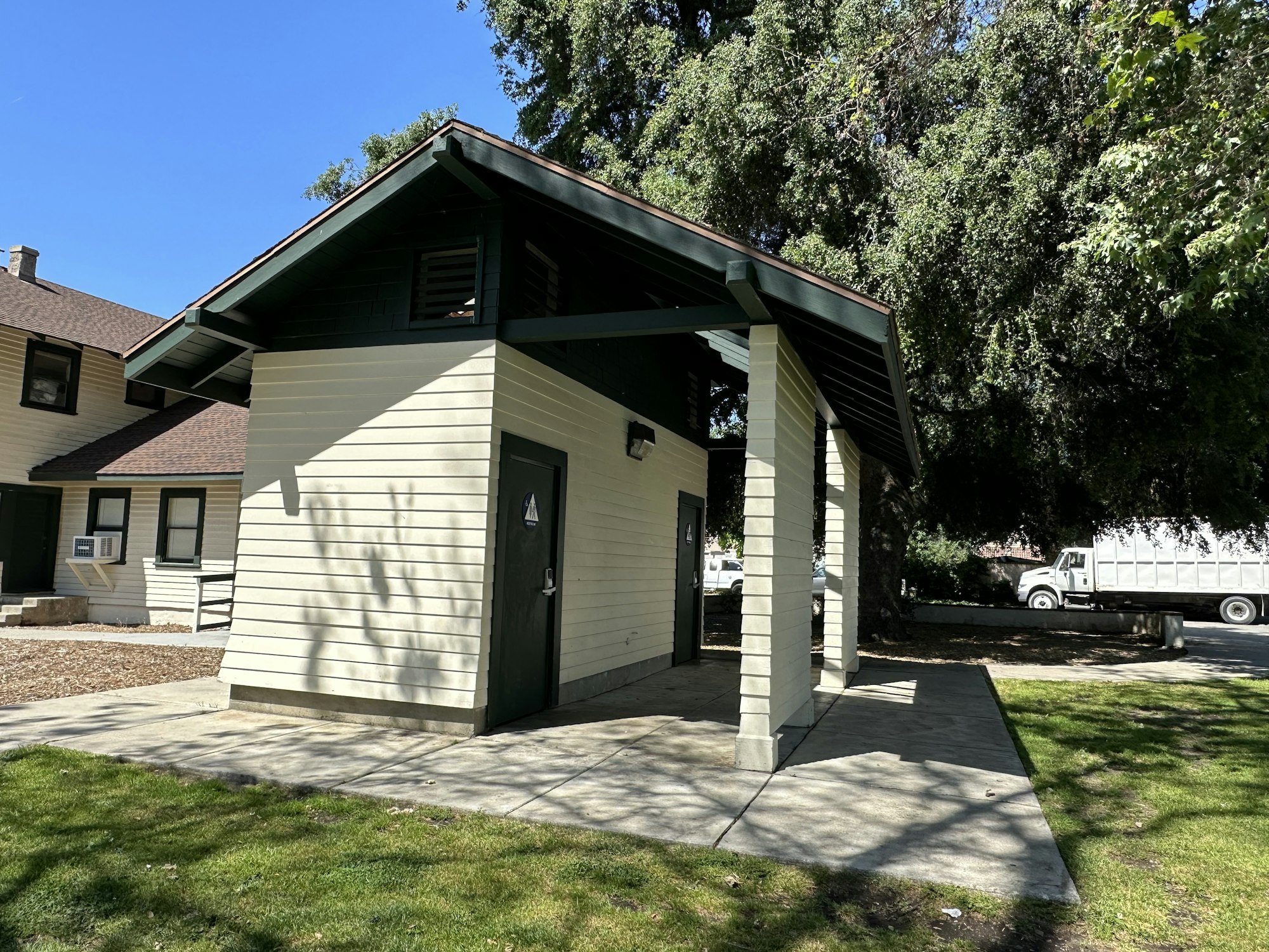A small building with a restroom sign, surrounded by grass and trees, next to a larger building and a parked truck.