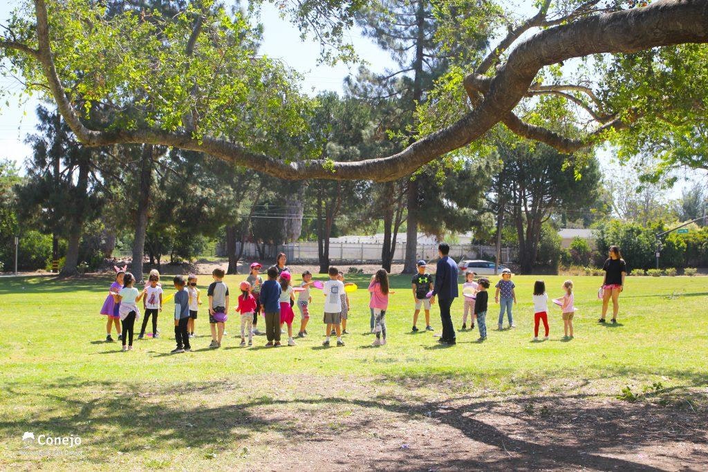 A group of children and two adults are playing in a park under a large tree.