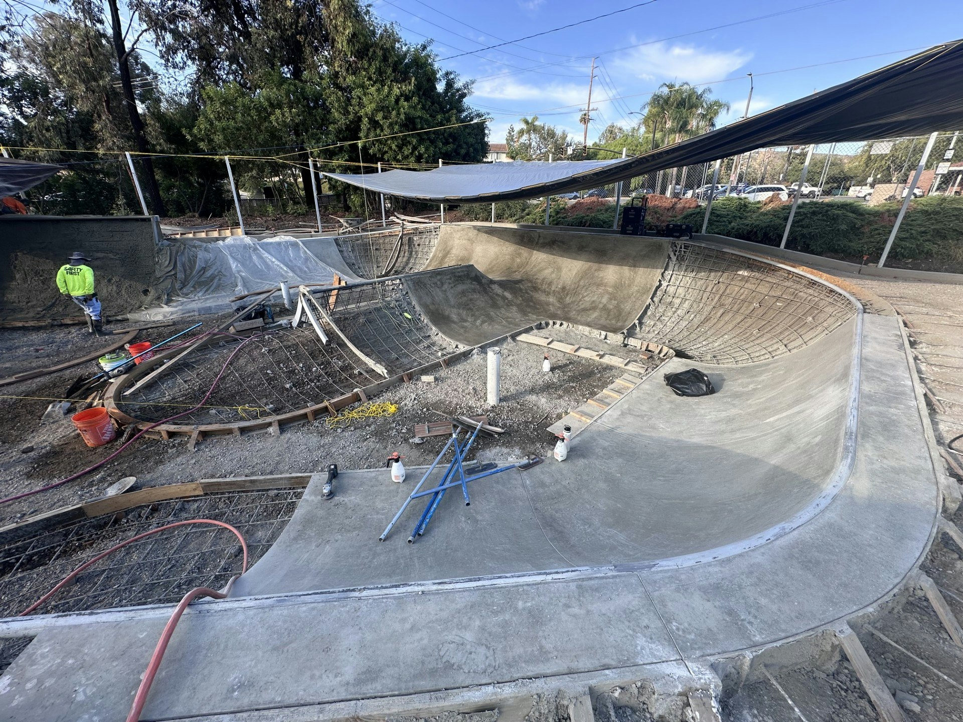 A construction site for a skate park, featuring unfinished concrete ramps and a worker in the background.