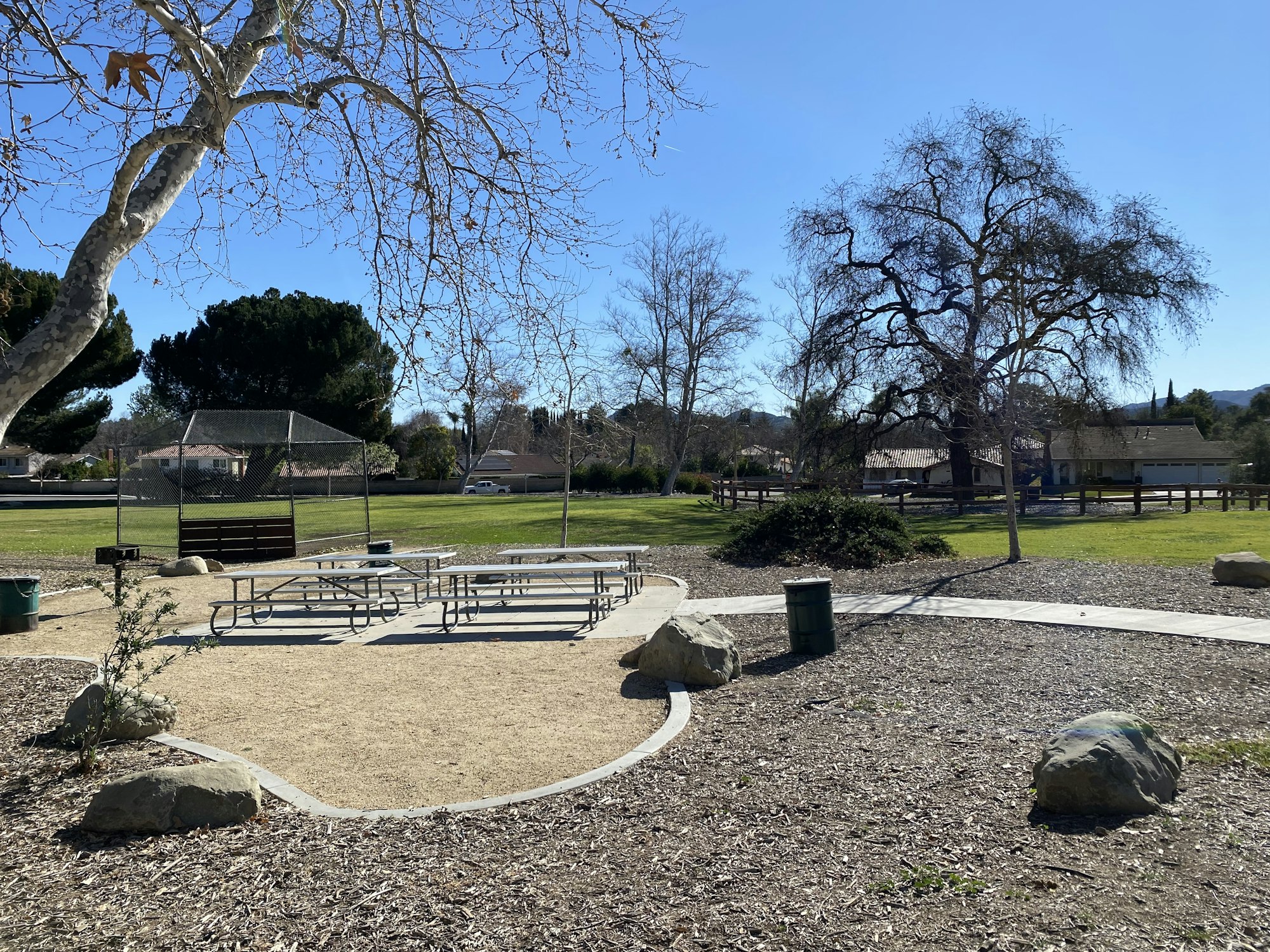 Park with picnic tables, trees, a grill, and a baseball backstop on a sunny day.