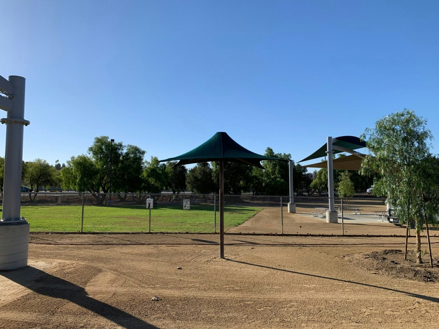 A fenced park area with trees, shade structures, and a paved path under a clear blue sky.