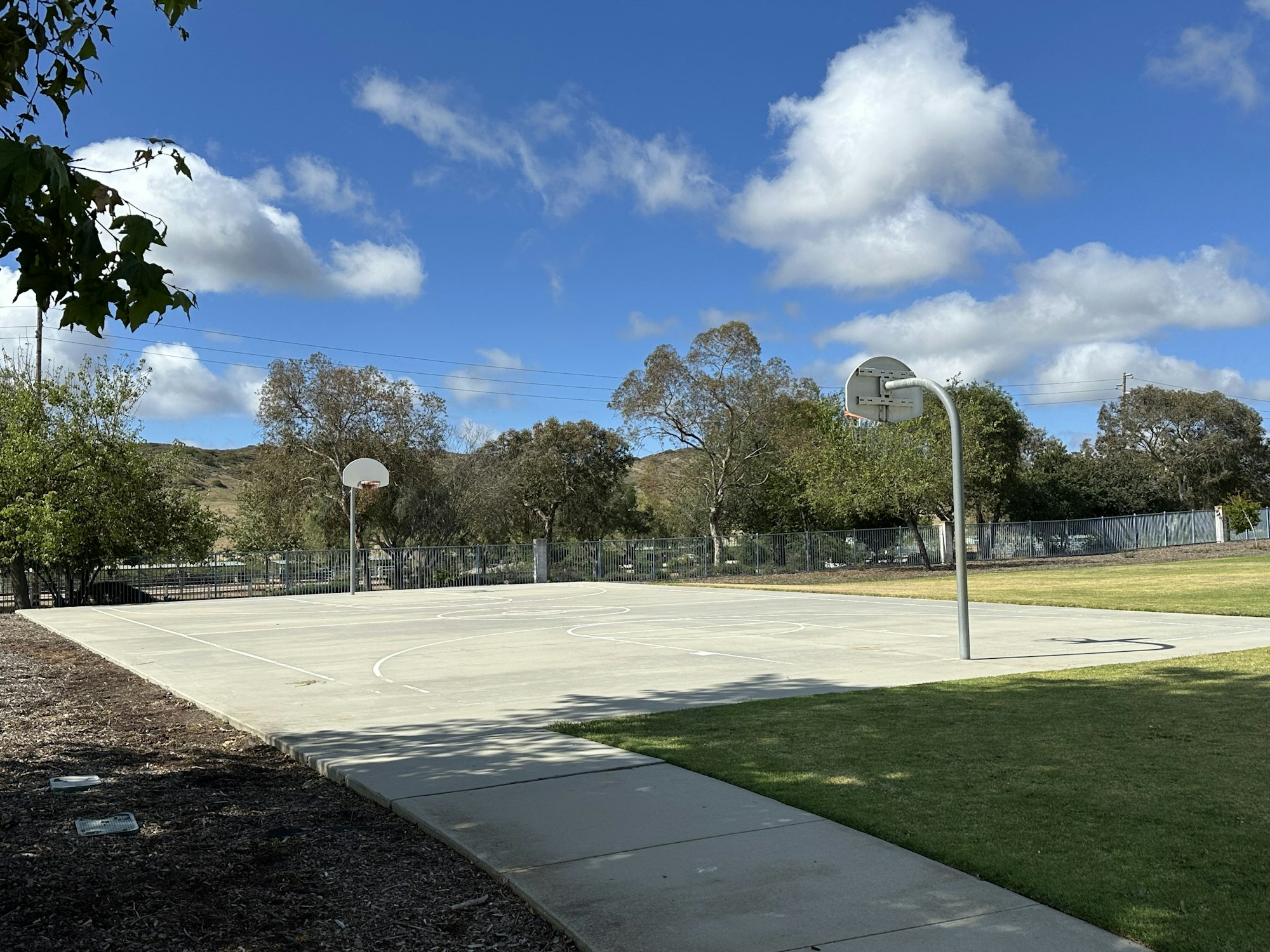 A basketball court under a blue sky with clouds, surrounded by trees and a fence.