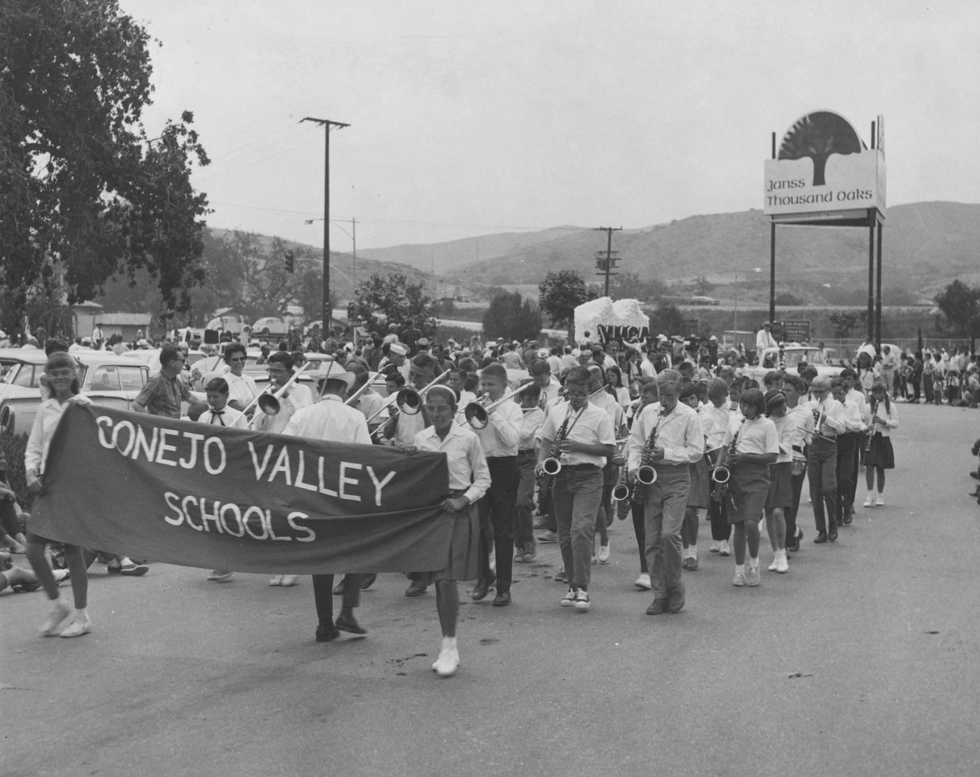 A marching band from Conejo Valley Schools parades holding a banner, with people watching, in black and white, mid-20th century setting.