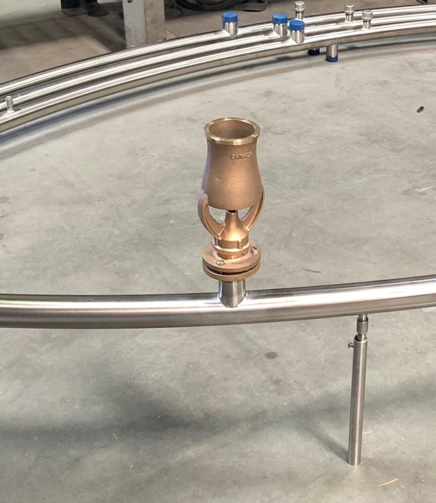 A copper nozzle mounted on a circular stainless steel pipe structure on a concrete floor.