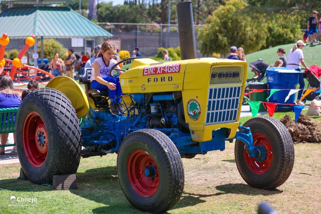 A child sits on a vintage Ford tractor at an outdoor event with people and colorful decorations in the background.