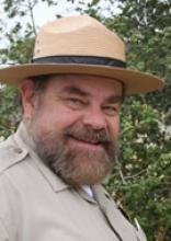 A smiling person wearing a hat with a green, leafy background.