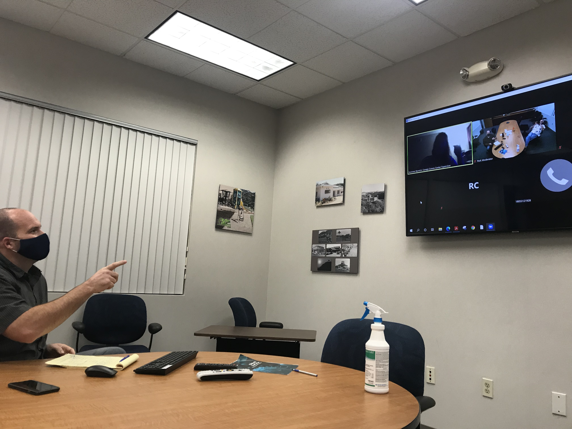A person wearing a mask is in a conference room, participating in a video call displayed on a wall-mounted screen.