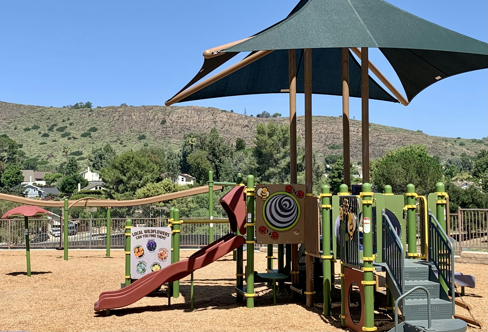 Playground with slides and shade canopy, surrounded by greenery and hills in the background.