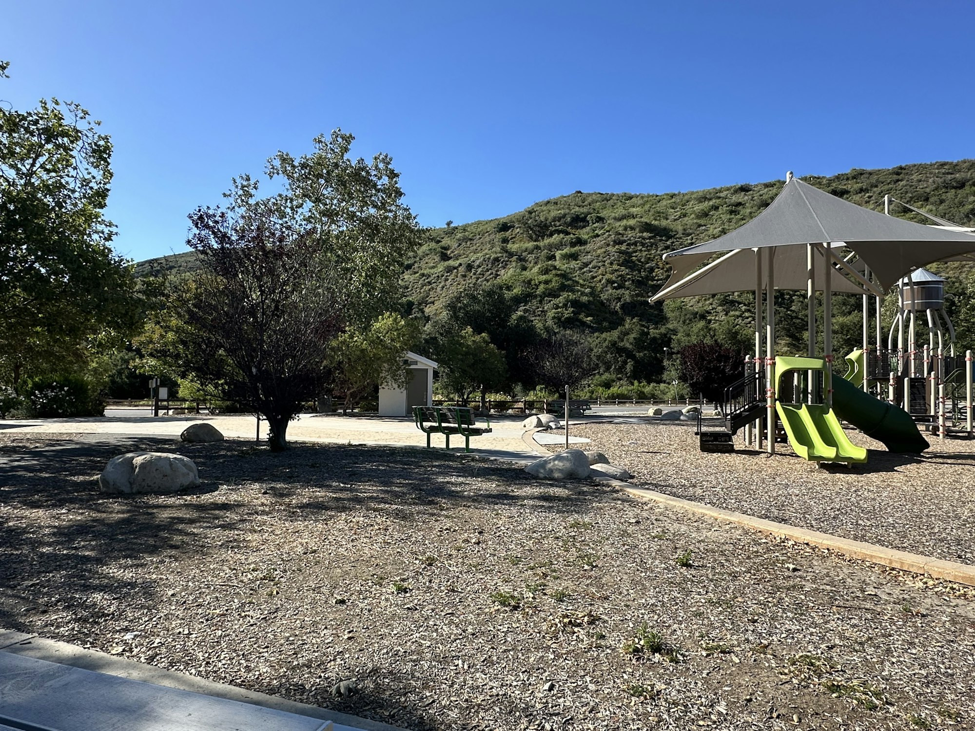 A sunny playground featuring a green slide, shaded structure, and surrounding greenery and rocky terrain.