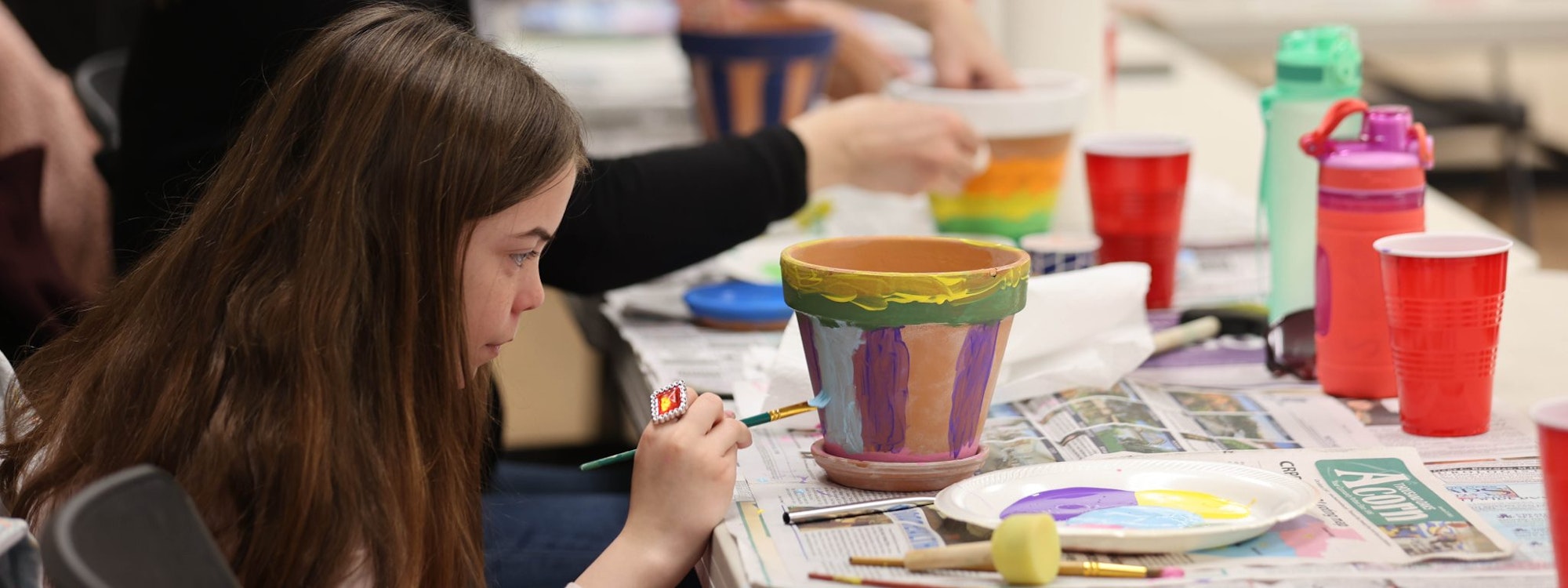 A child painting a flower pot at a table with art supplies, cups, and a newspaper.