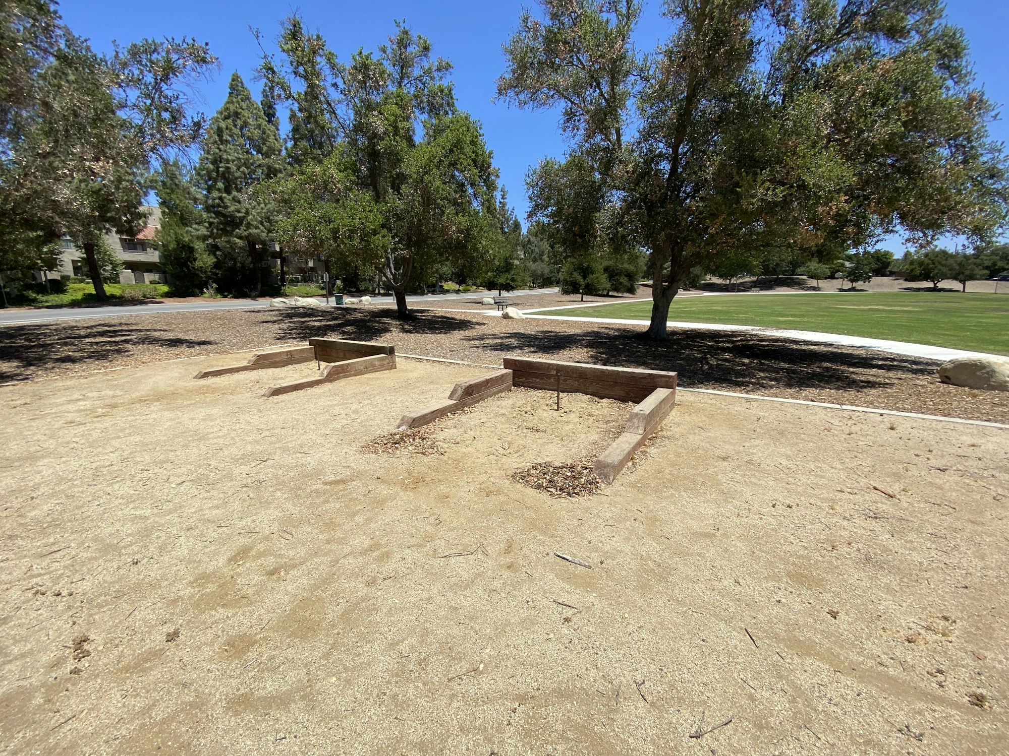 Outdoor horseshoe pits surrounded by trees and a grassy field under a clear blue sky.