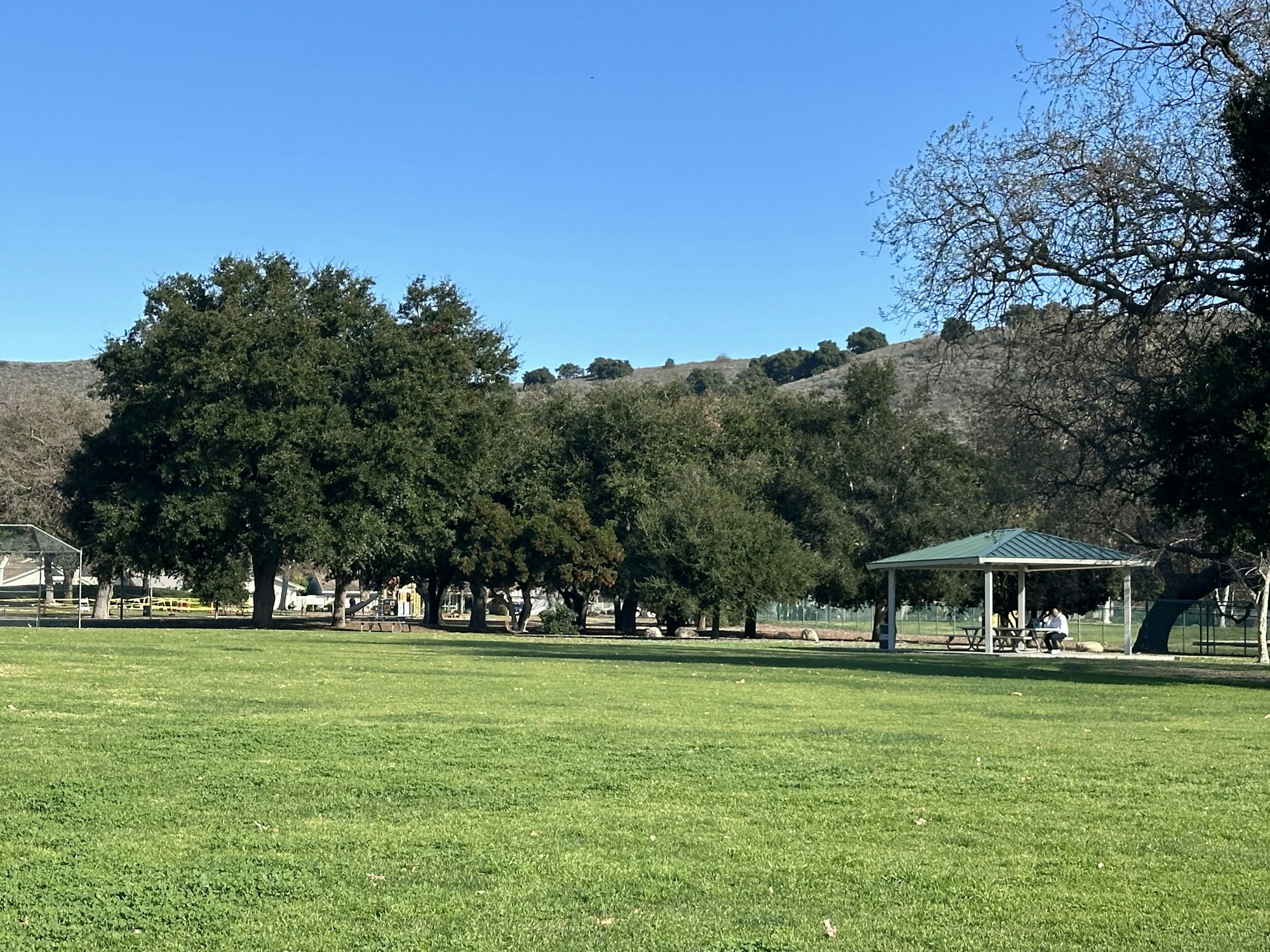 A park with green grass, trees, a small pavilion, and hills in the background under a clear blue sky.