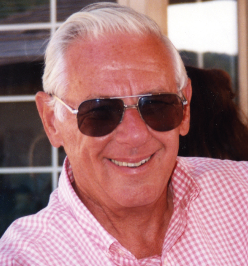 An older man wearing sunglasses and a pink checkered shirt, smiling outdoors.