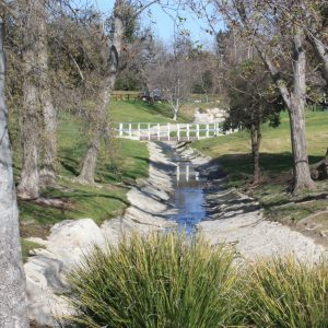 A small stream with rocks, grass, and trees on both sides. A white fence crosses the stream in the distance.