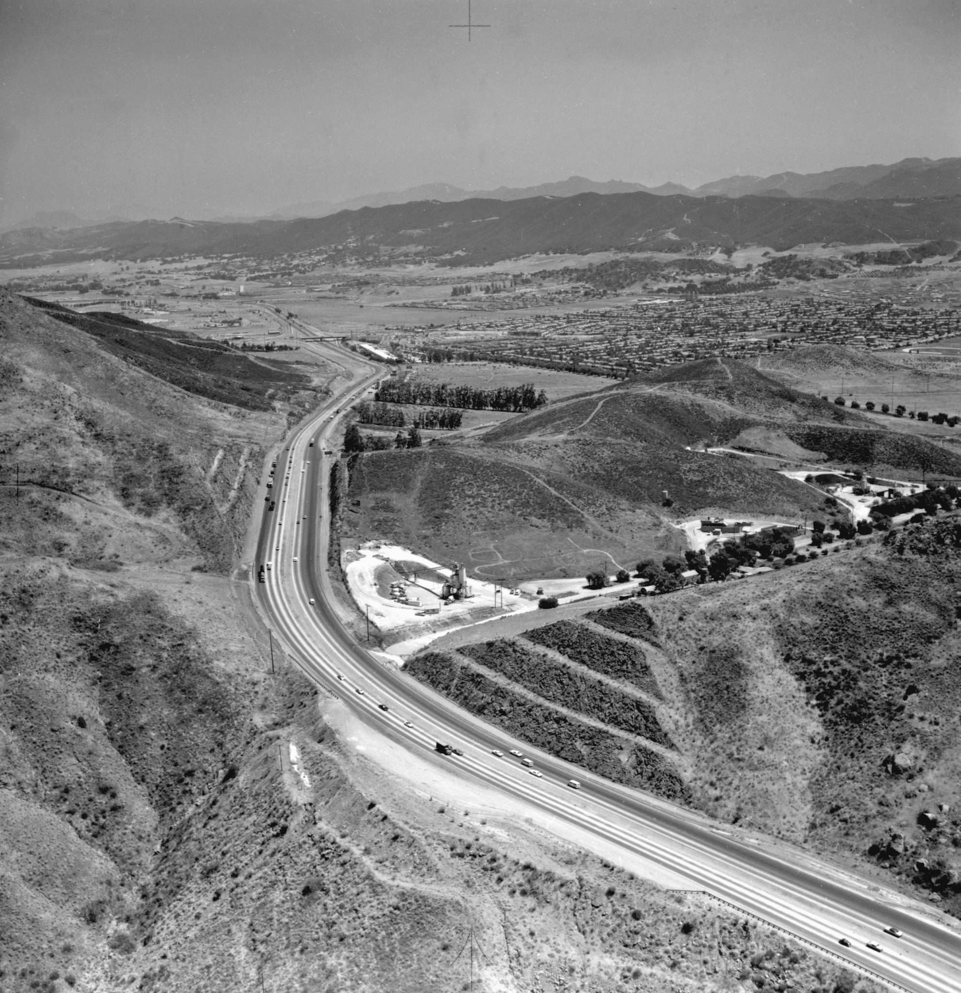 Aerial view of a highway curving through hills, with sparse traffic, buildings, and distant mountains in the background.